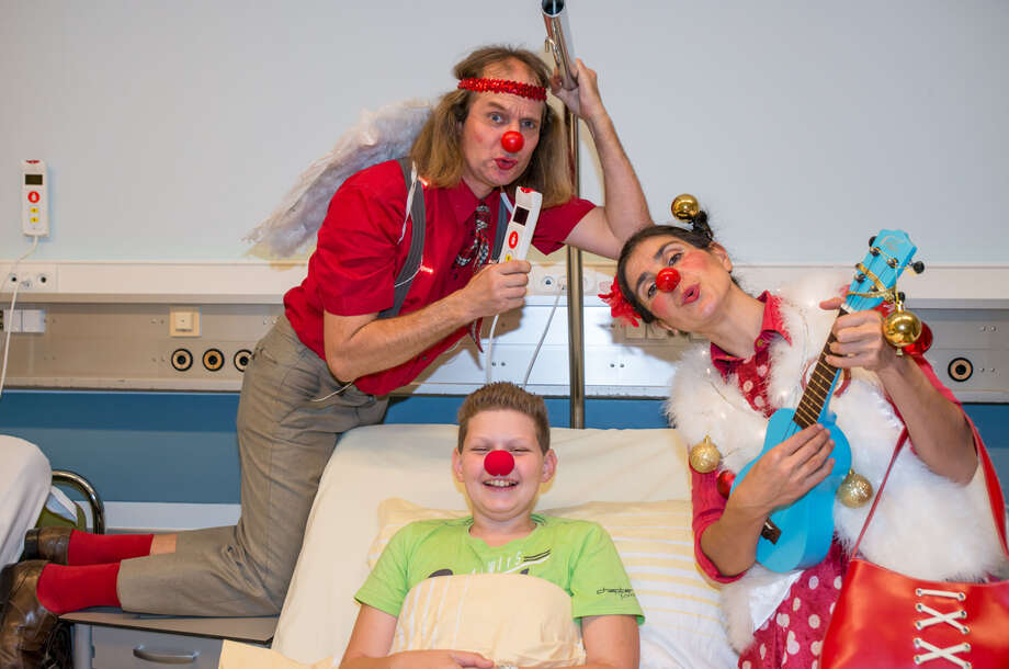 lachender Junge mit roter Schaumstoffnase im Spitalsbett, rechts neben ihm steht eine Clownin mit Ukulele, Clown mit Engelsflügeeln hält sich oben am Bett fest und singt