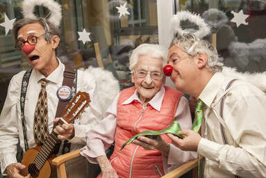 Seniorin sitzt zwischen zwei Clowns, die Engelsflügerl und einen Heiligenschein tragen, sie singen gemeinsam