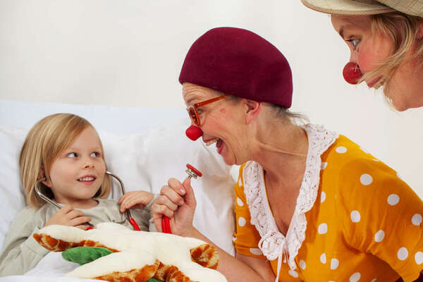 Zwei ROTE NASEN Clown:innen bringen ein Kind im Krankenhausbett zum Lachen.