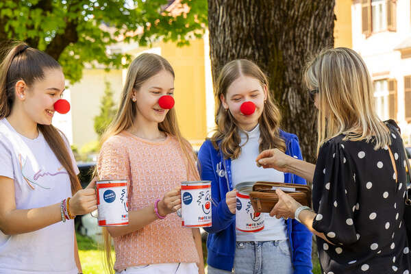 Drei junge Menschen mit roten Clownnasen sammeln im Freien Spenden in ROTE NASEN Spendenboxen, eine Frau wirft Geld hinein.