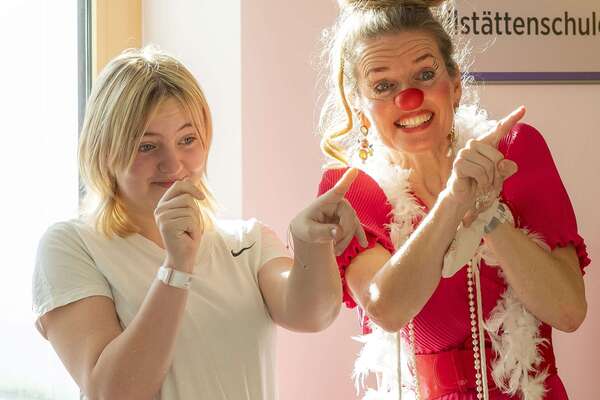 Ein ROTE NASEN Clown in auffälligem roten Kleid mit Federboa steht lachend neben einem Mädchen, beide machen eine witzige Geste.