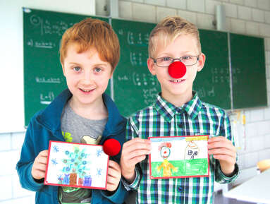Zwei Schulkinder stehen vor einer Tafel und halten selbstgezeichnete Grußkarten, einer der Jungen trägt eine ROTE NASE