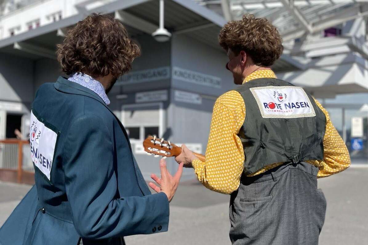 Zwei ROTE NASEN Clowns gehen mit Instrument vor einem Krankenhausgebäude, auf ihren Jacken sind die Logos der ROTE NASEN sichtbar.