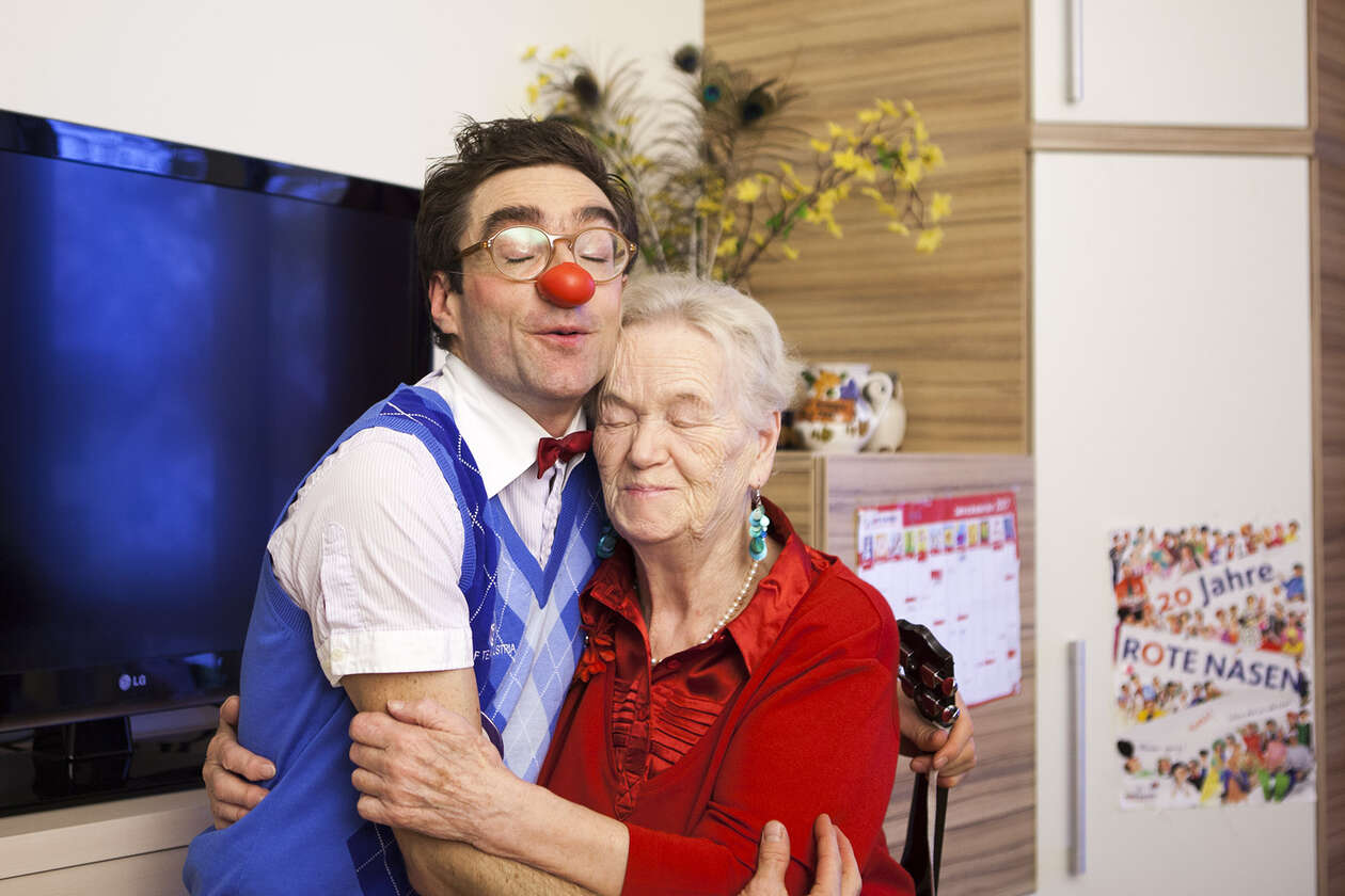 Clown und Seniorin nehmen sich innig in die Arme und haben die Augen dabei geschlossen
