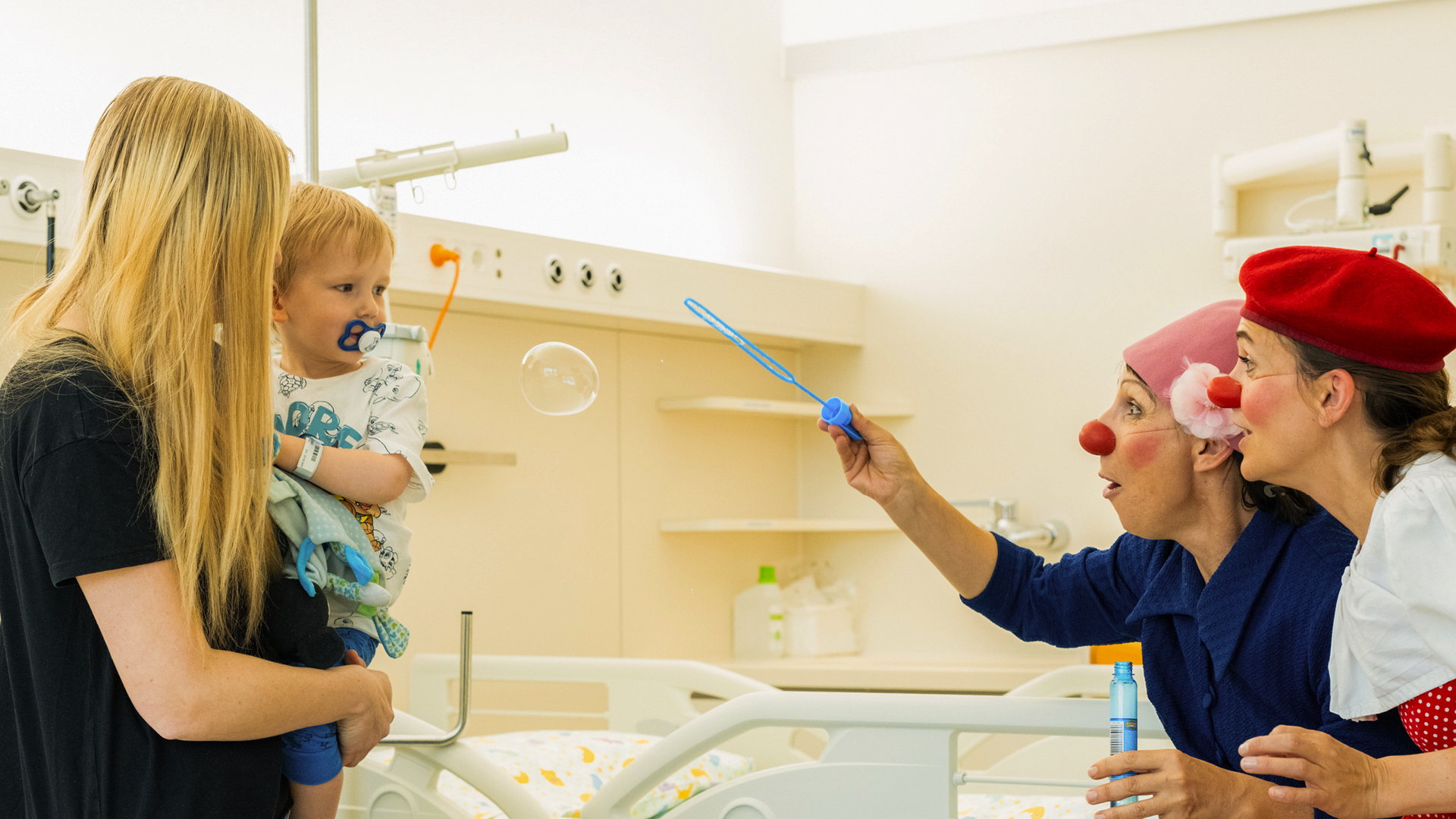 Zwei ROTE NASEN Clown:innen pusten Seifenblasen für ein kleines Kind, das auf dem Arm seiner Mutter im Krankenhauszimmer sitzt.
