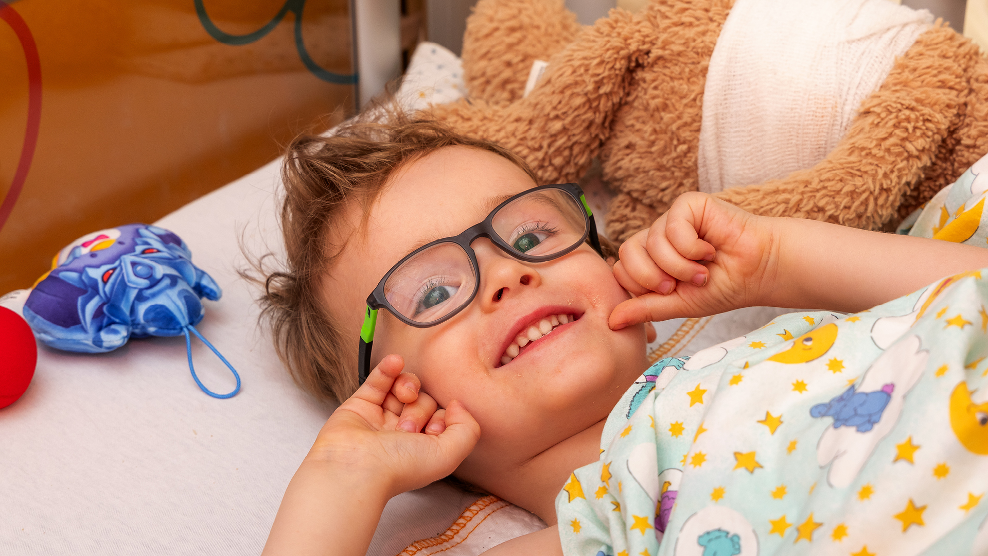 Ein lachendes Kind mit Brille liegt im Krankenhausbett, kuschelt mit einem Teddybären und schaut fröhlich in die Kamera.