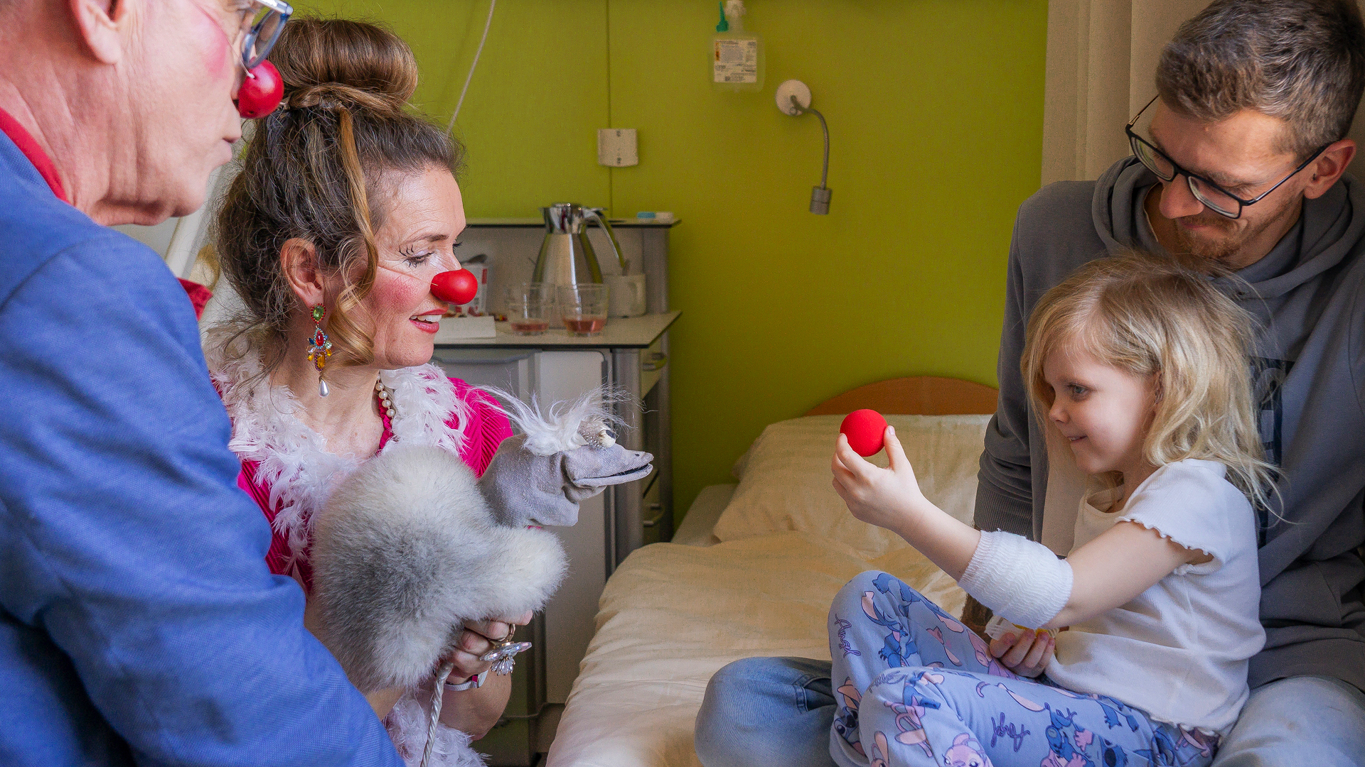 Zwei ROTE NASEN Clowns besuchen ein Mädchen im Krankenhaus, das mit einem roten Clownnasen-Ball spielt, während ein Erwachsener daneben sitzt.