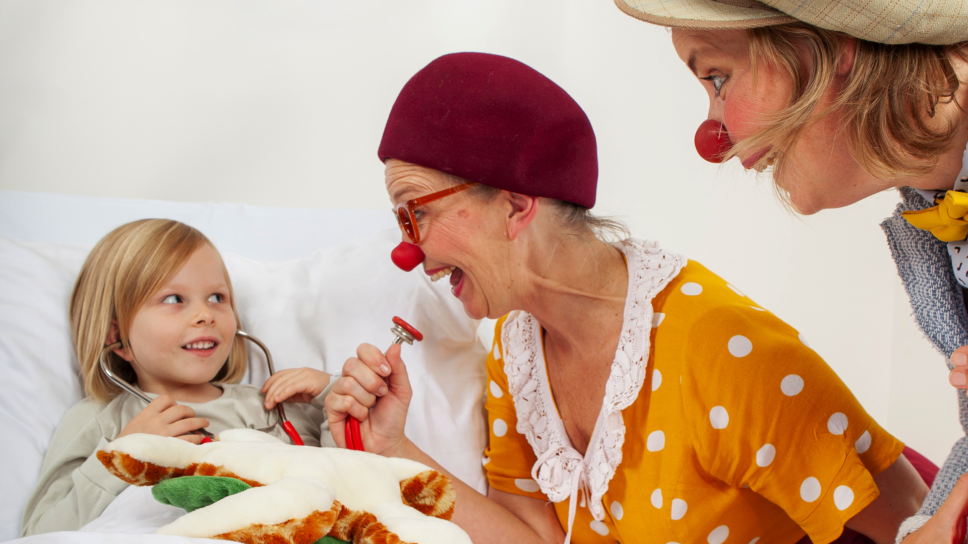 Zwei ROTE NASEN Clown:innen bringen ein Kind im Krankenhausbett zum Lachen.