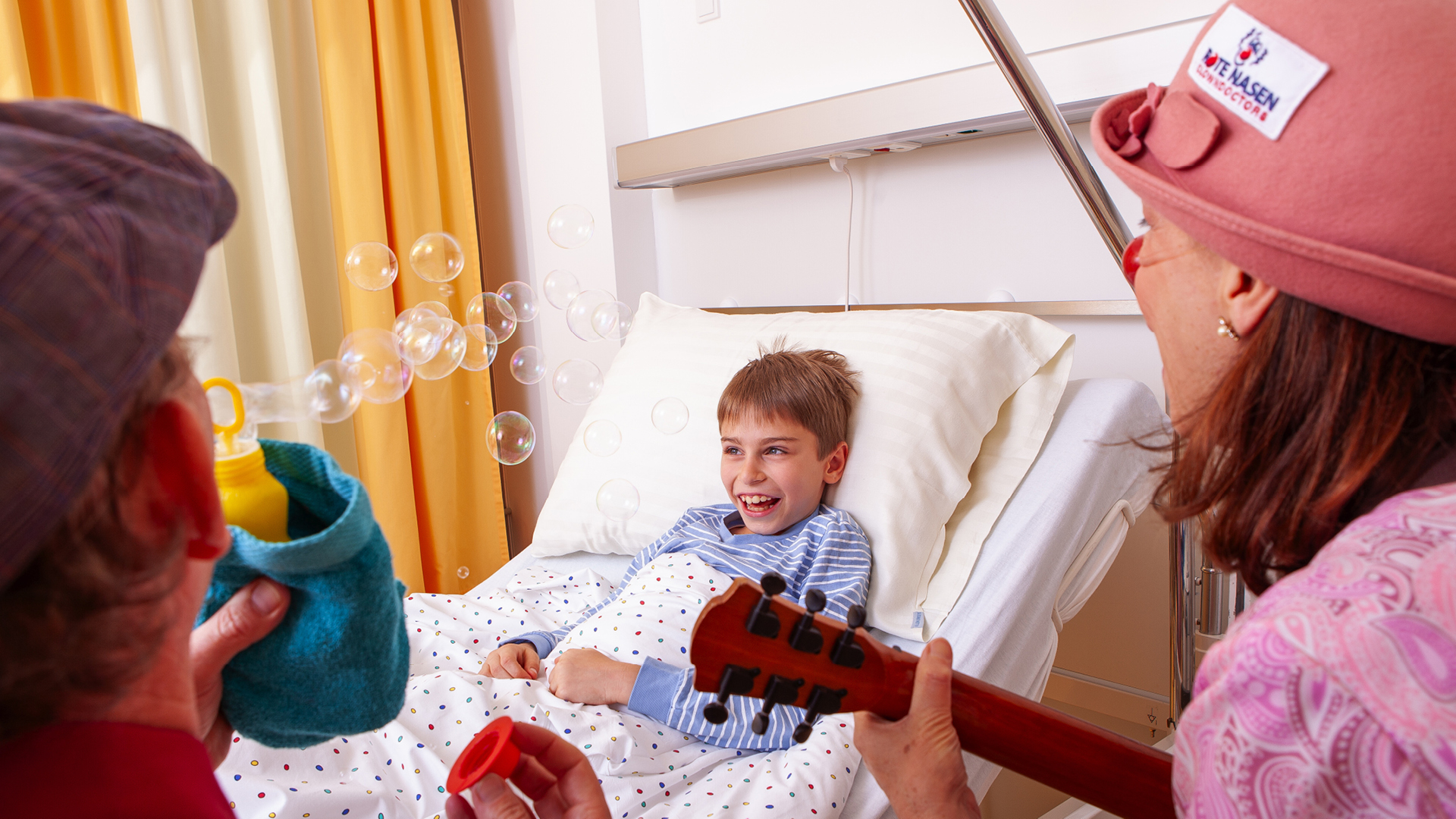 Zwei ROTE NASEN Clown:innen spielen Musik und pusten Seifenblasen für ein lachendes Kind im Krankenhausbett.