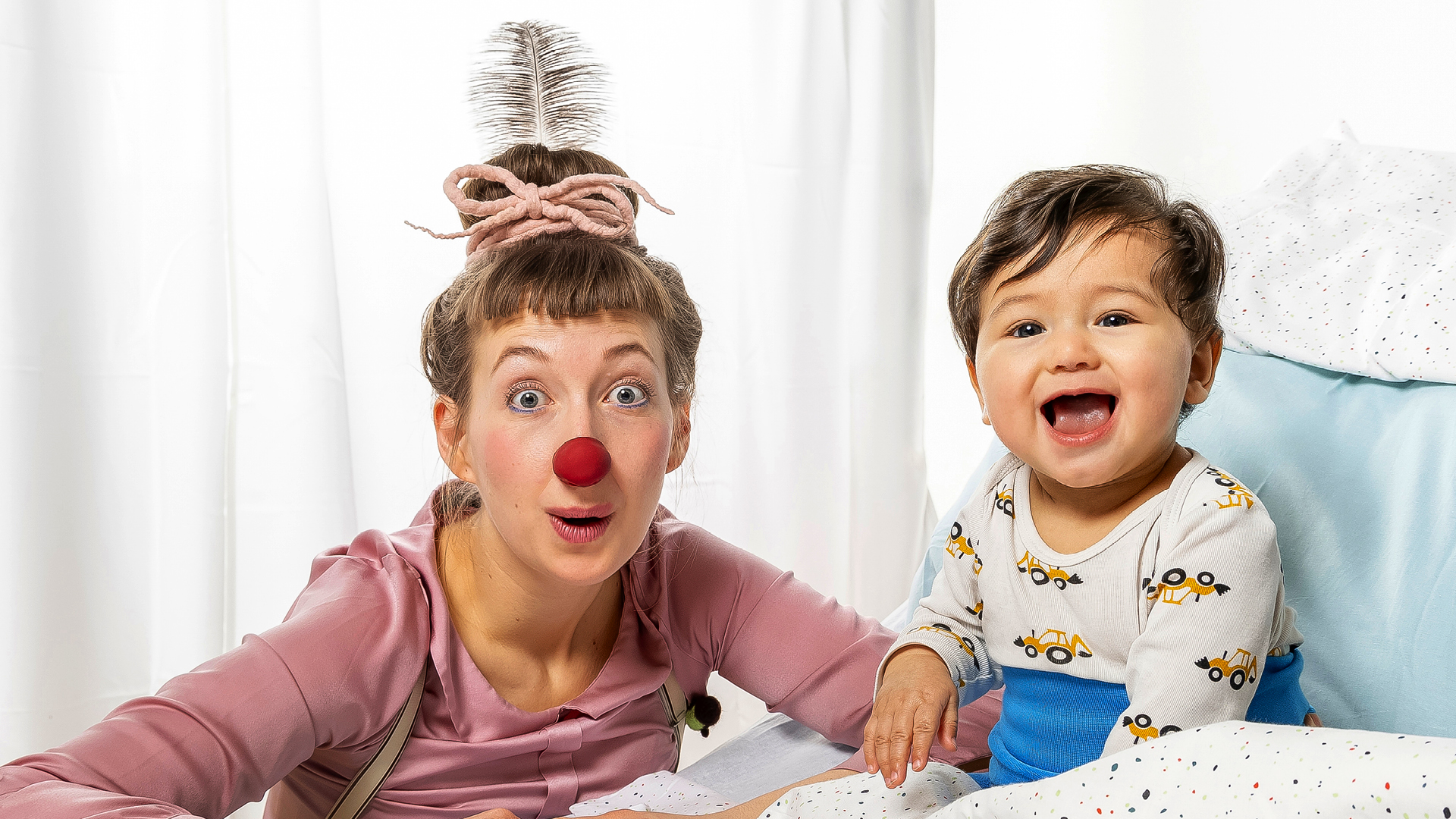 Eine ROTE NASEN Clownin macht lustige Grimassen neben einem lachenden Kind im Krankenhausbett.