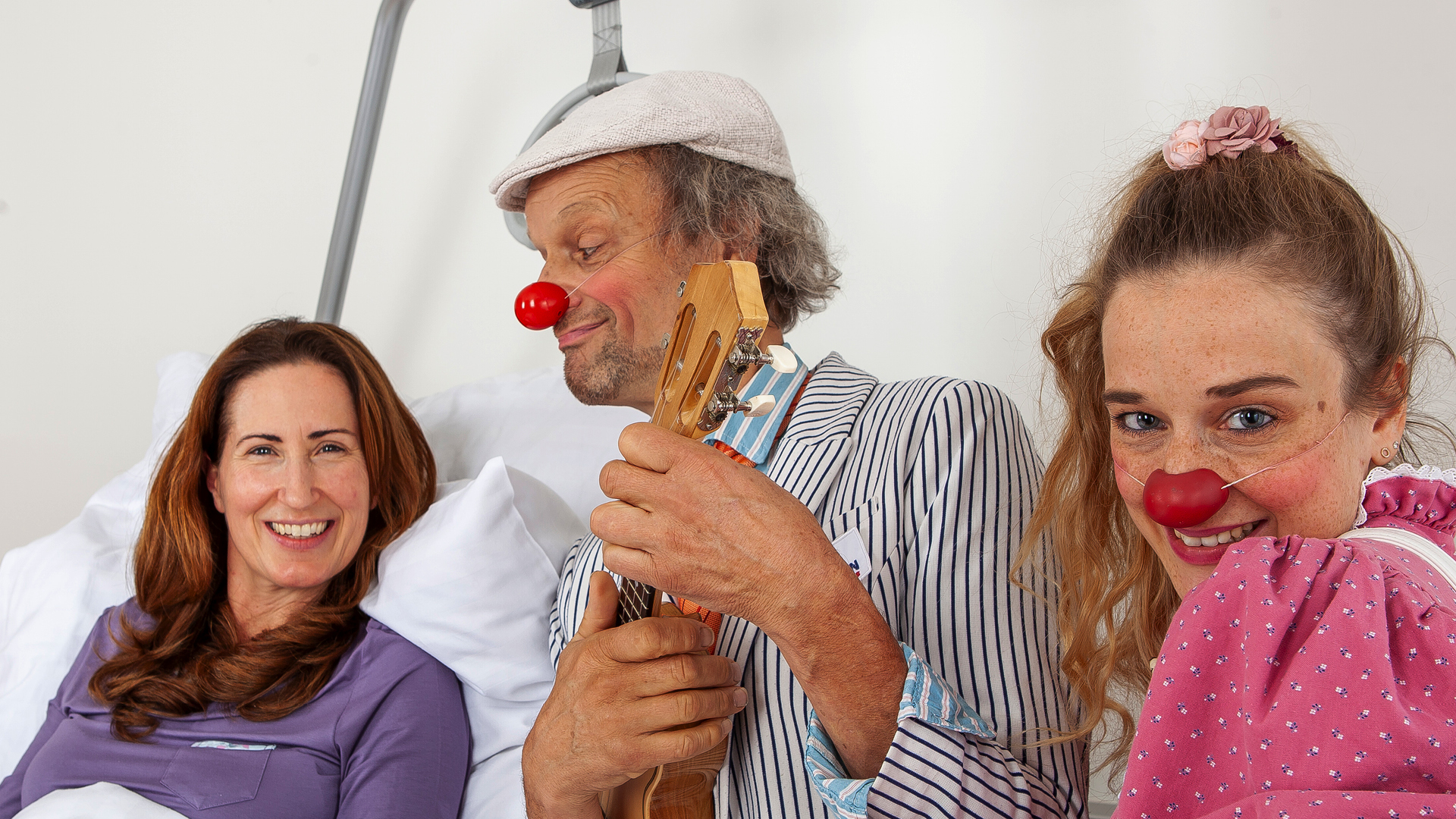 Zwei ROTE NASEN Clown:innen bringen eine Frau im Krankenhausbett zum Lächeln – der Clown spielt Ukulele, die Clownin schaut lächelnd in die Kamera.