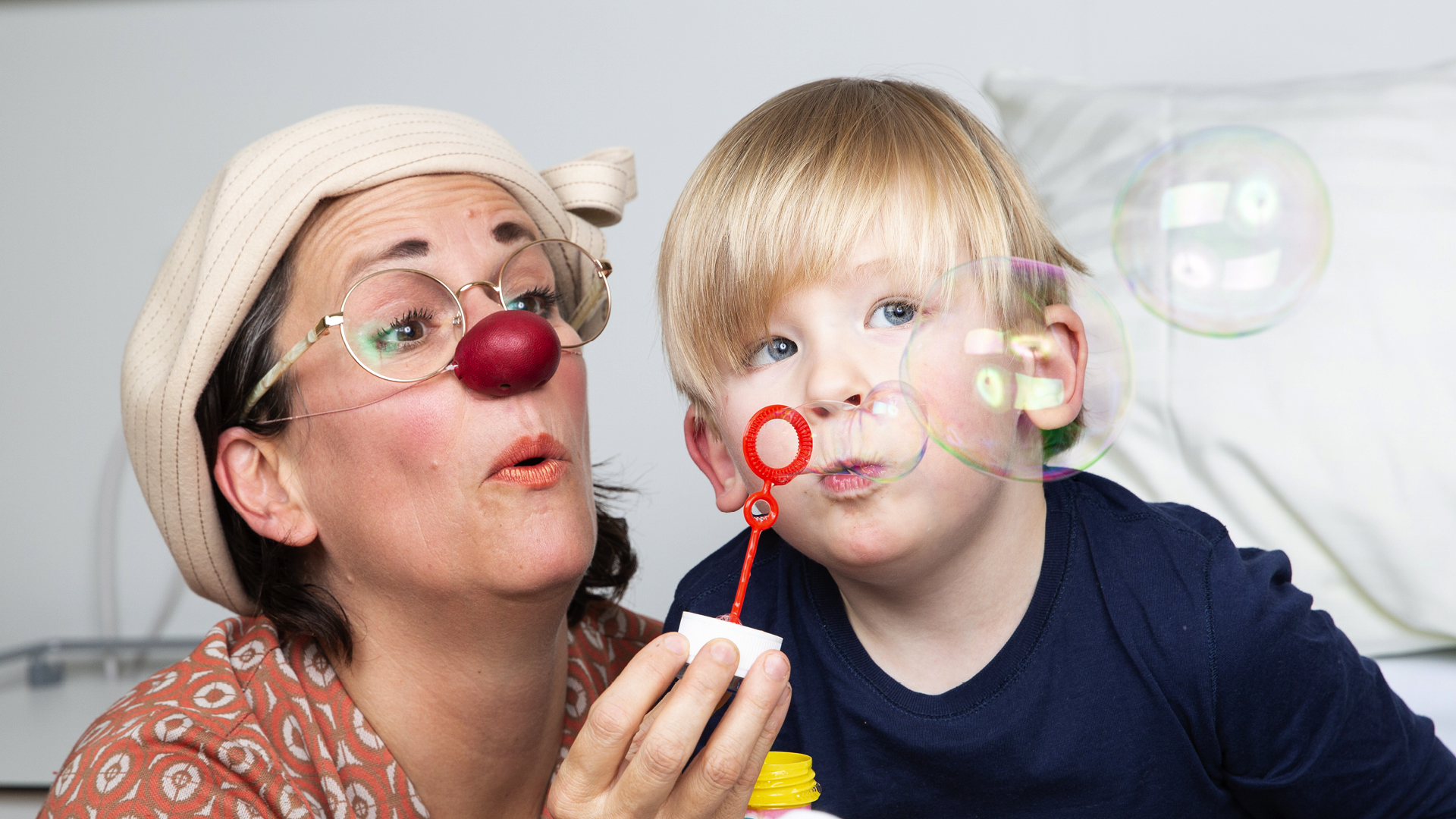 Eine ROTE NASEN Clownin pustet gemeinsam mit einem kleinen Kind Seifenblasen im Krankenhauszimmer.