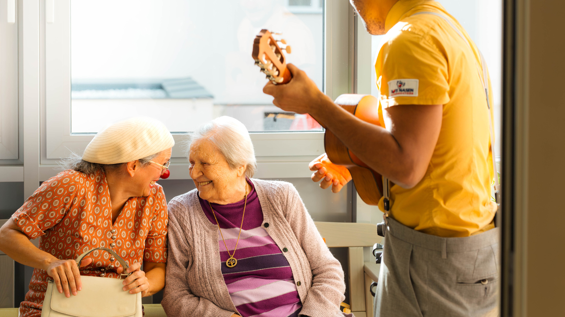 Zwei ROTE NASEN Clowns besuchen eine ältere Frau, die lächelnd im Stuhl sitzt, während einer der Clowns Ukulele spielt.