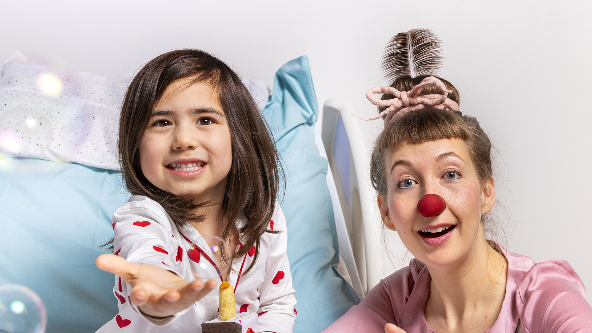 Eine ROTE NASEN Clownin spielt mit einem Kind im Krankenhausbett, beide lachen und beobachten Seifenblasen.