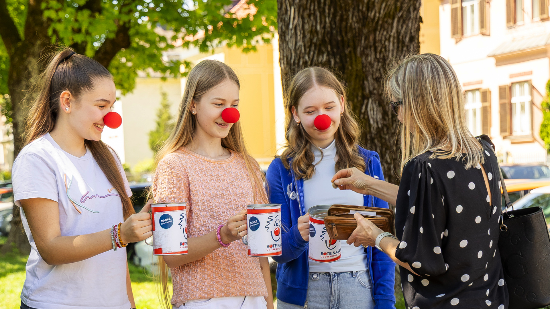 Drei junge Menschen mit roten Clownnasen sammeln im Freien Spenden in ROTE NASEN Spendenboxen, eine Frau wirft Geld hinein.