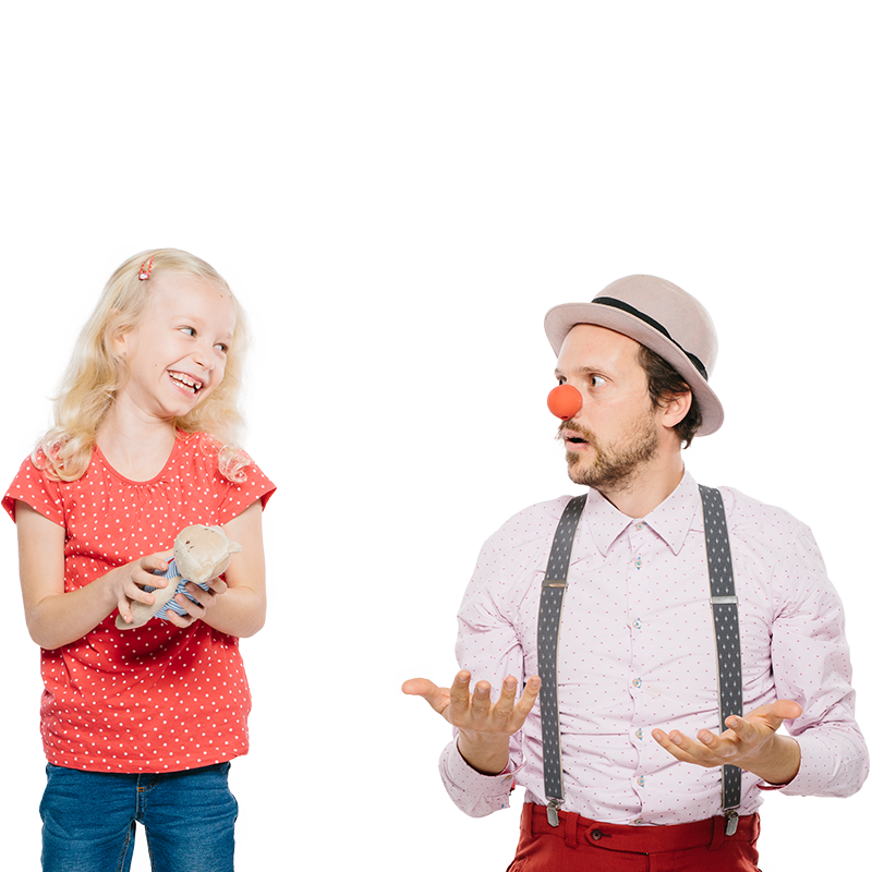 Mödchen mit Teddybär lächelt schelmisch Clown neben ihr an der fragend die Hände und Schultern in die Luft hebt