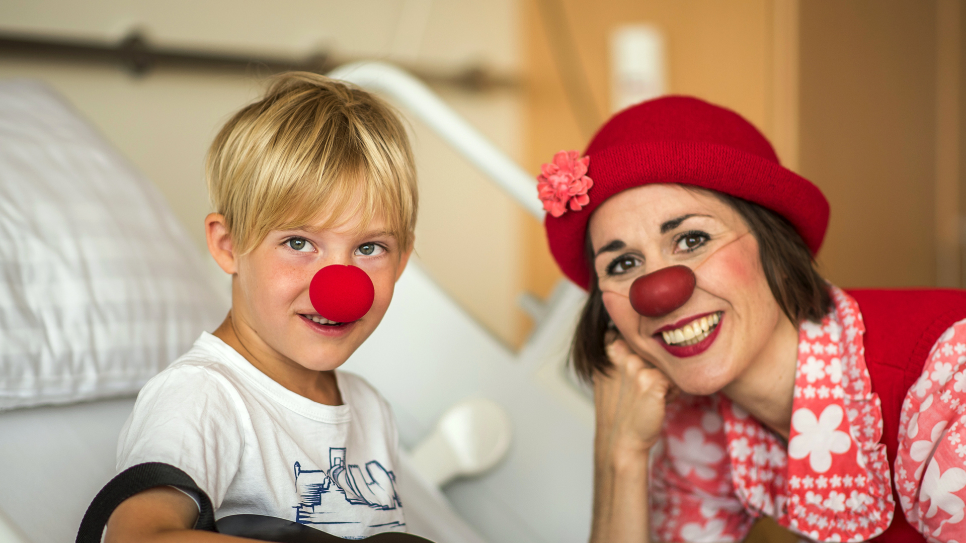 Ein Kind und eine ROTE NASEN Clownin mit roter Nase und rotem Hut sitzen lachend im Krankenhauszimmer.