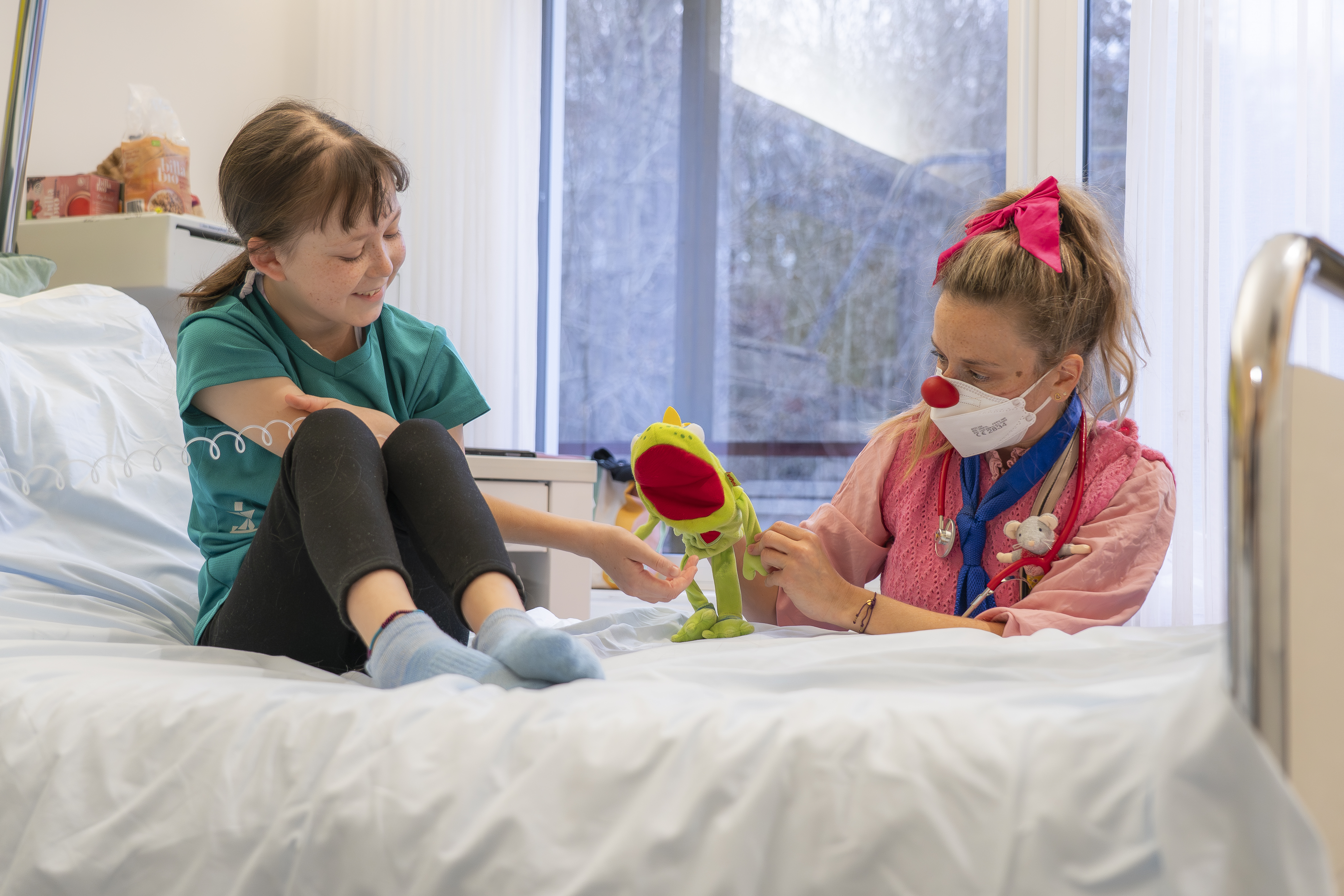ROTE NASEN Clownin spielt mit einer grünen Handpuppe für ein lachendes Mädchen im Krankenhausbett.