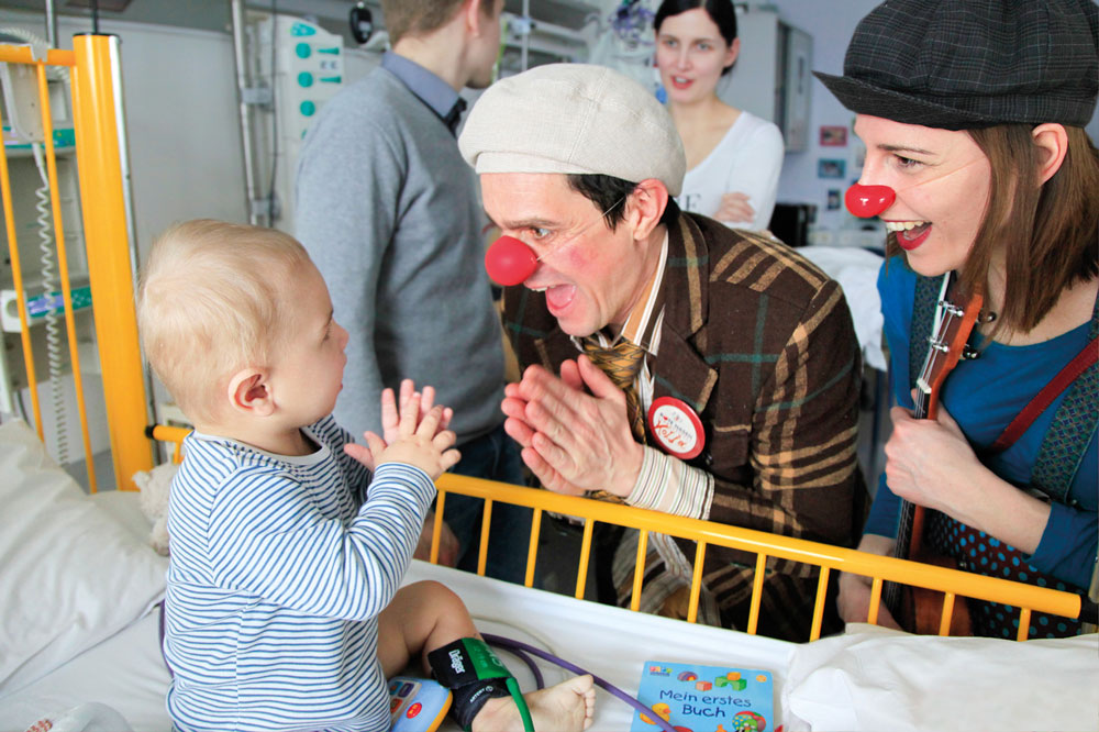 Schenkst du mir auch morgen ein Lachen? Clownin und Clown interagieren mit Kleinkind im Krankenhausgitterbett, der kleine Junge klatscht