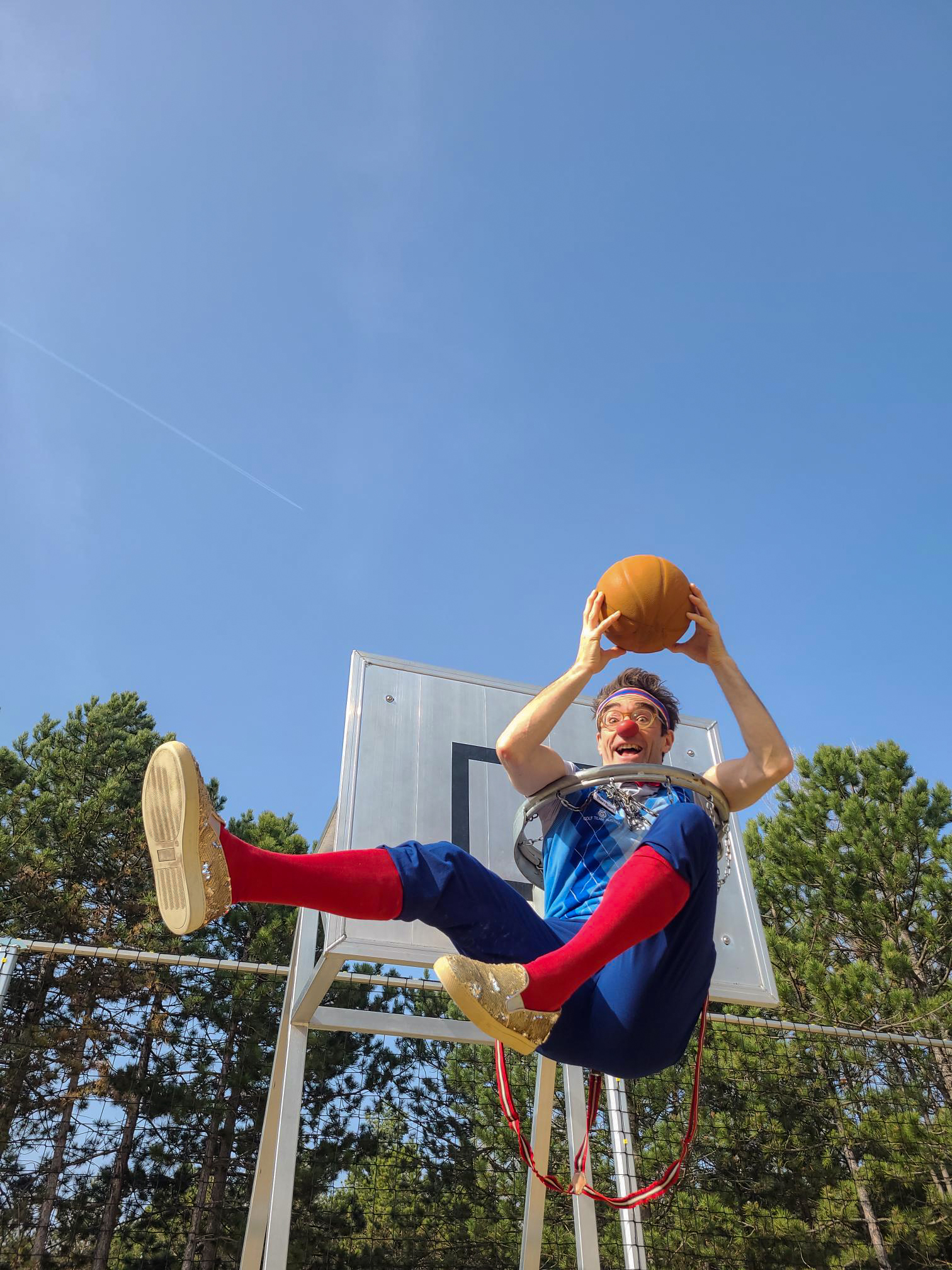 Clown Bernhart sitzt in einem Basketballkorb und hält einen Ball.