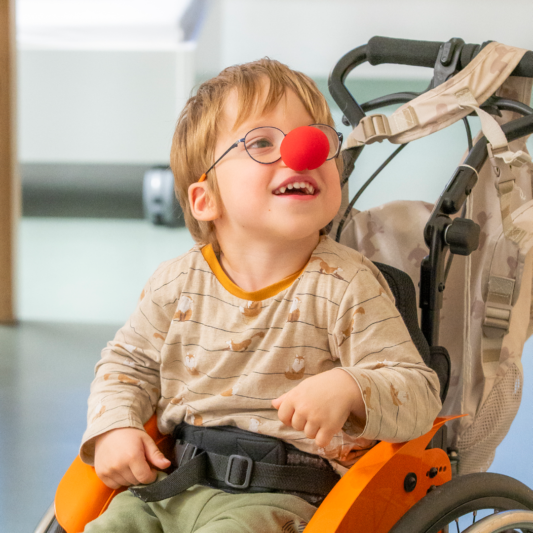 Junge in Rollstuhl mit einer roten Clownnase auf seiner Nase.