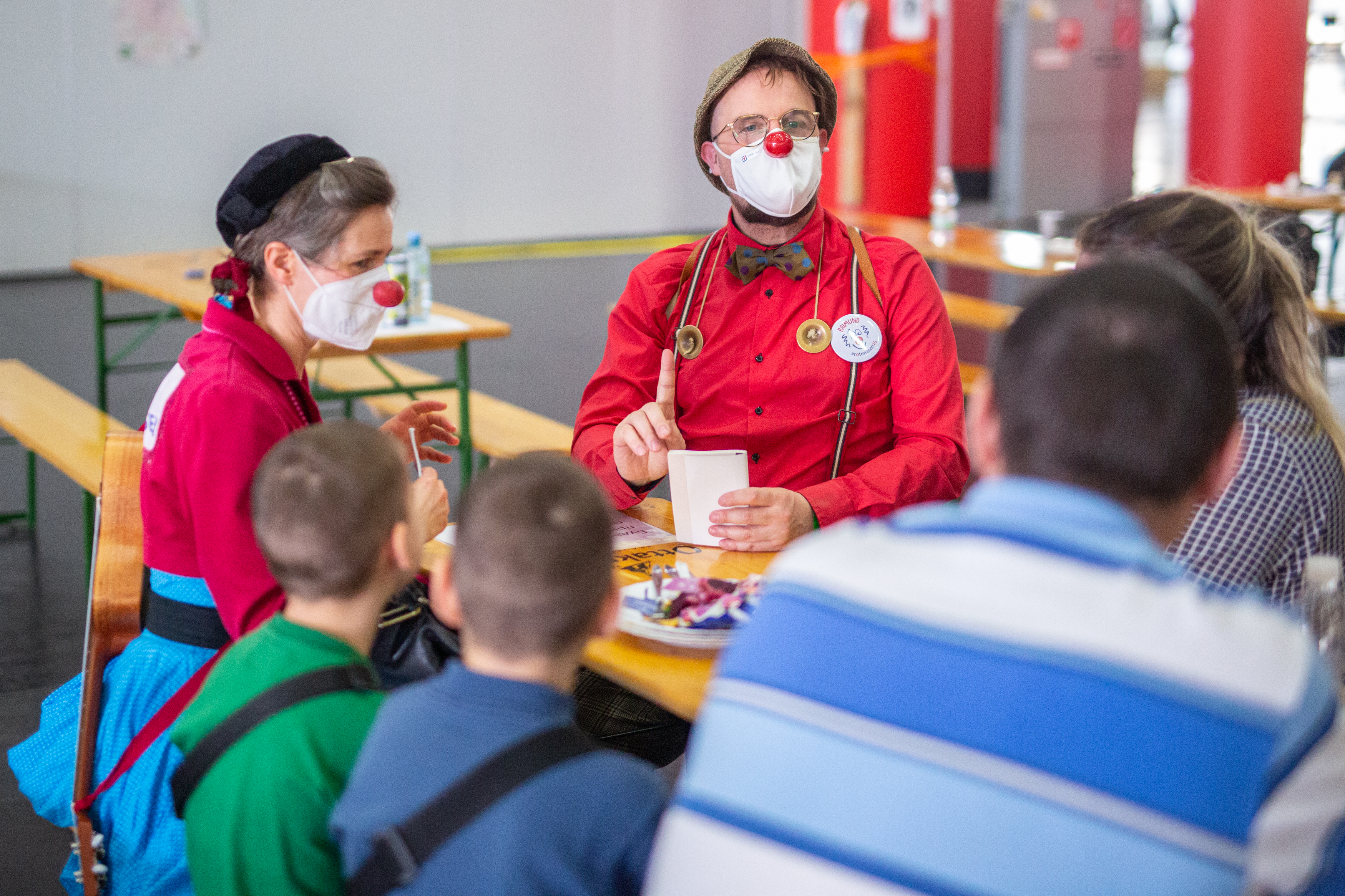 ROTE NASEN-Clowns Irmi und Raimund sitzen an einem Tisch mit zwei Erwachsenen und zwei Kindern.
