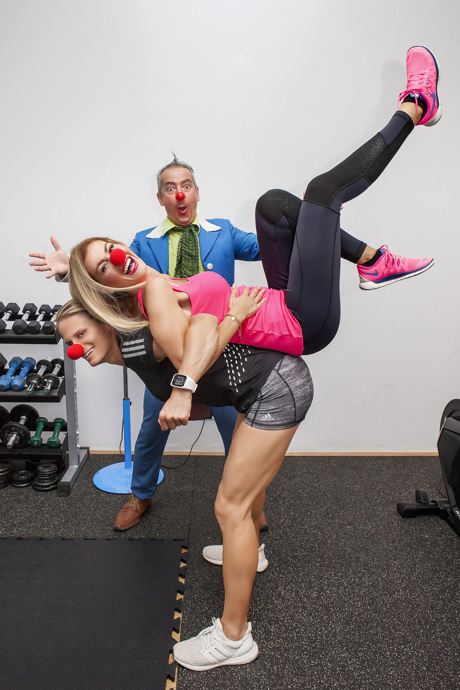 Eine Frau steht nach vorne gebeugt, die zweite Frau liegt mit ihrem Rücken auf der ersten Frau und wir von ihr hochgehoben, dahinter posiert ein ROTE NASEN Clown