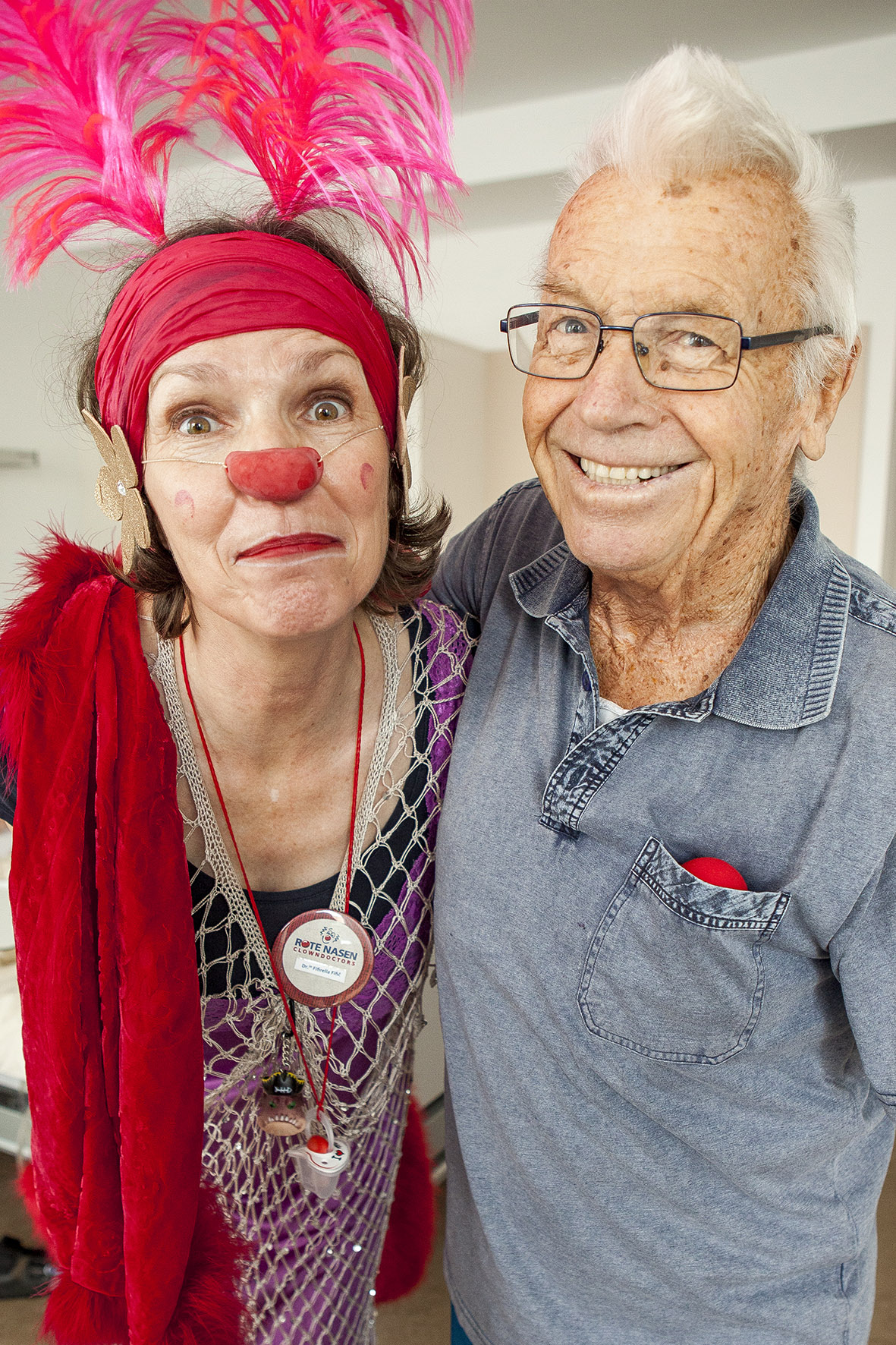 Eine Clownin mit Federn im Haar posiert gemeinsam mit einem Senior. Der Senior lacht herzlich in die Kamera.