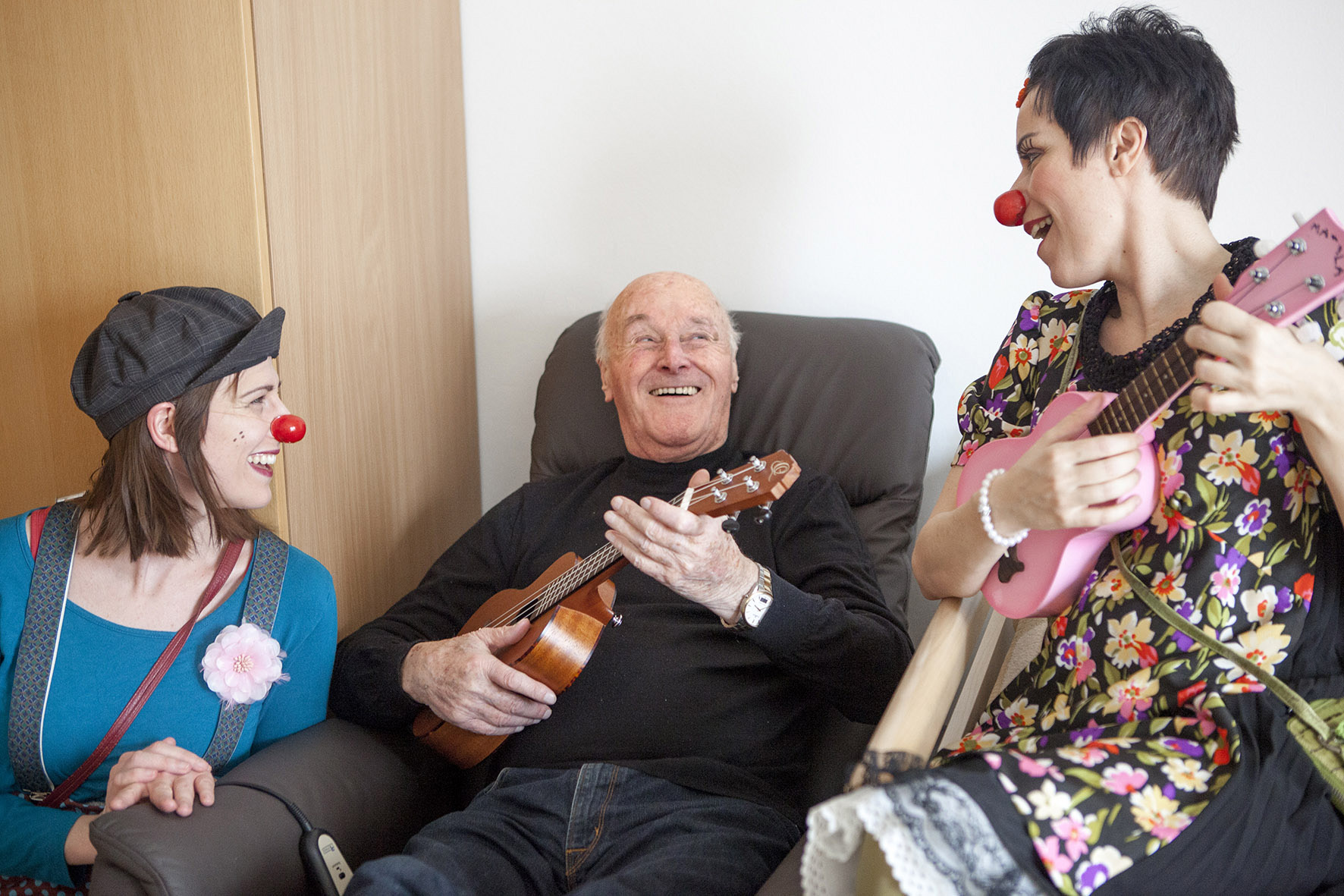 Zwei Clowninnen sitzen links und rechts von einem Senior im Stuhl, er hält eine Ukulele in der Hand und spielt, eine der Clowninnen spielt ebenfalls Ukulele und sie lachen sich an
