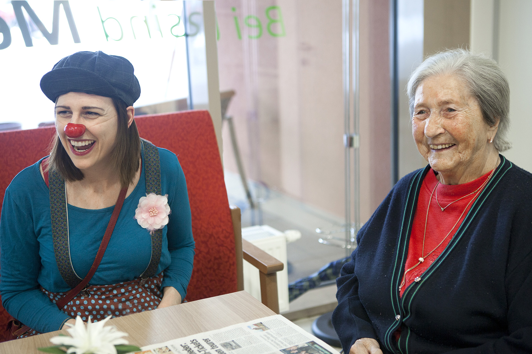 ROTE NASEN-Clownin Marie sitz neben einer älteren Dame und beide lachen herzhaft.