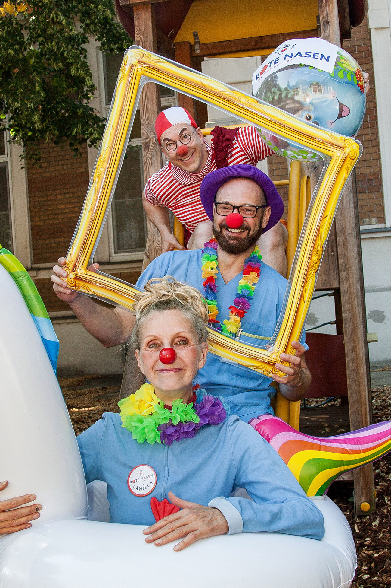 Clownin in einem Plastikeinhorn sitzt vor einem Mitarbeiter des Krankenhauses der einen goldenen Rahmen hält, dahinter steht ein weiterer Clown mit einem Plastikball