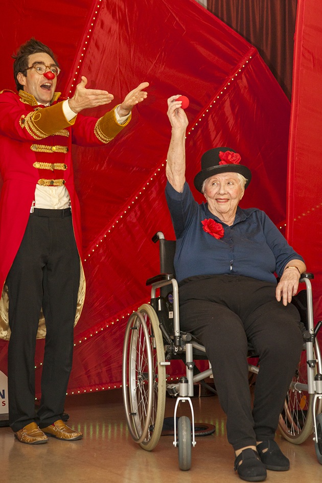 Clown präsentiert den Auftritt einer Seniorin im Rollstuhl, die lächelnd eine rote Nase in die Luft streckt