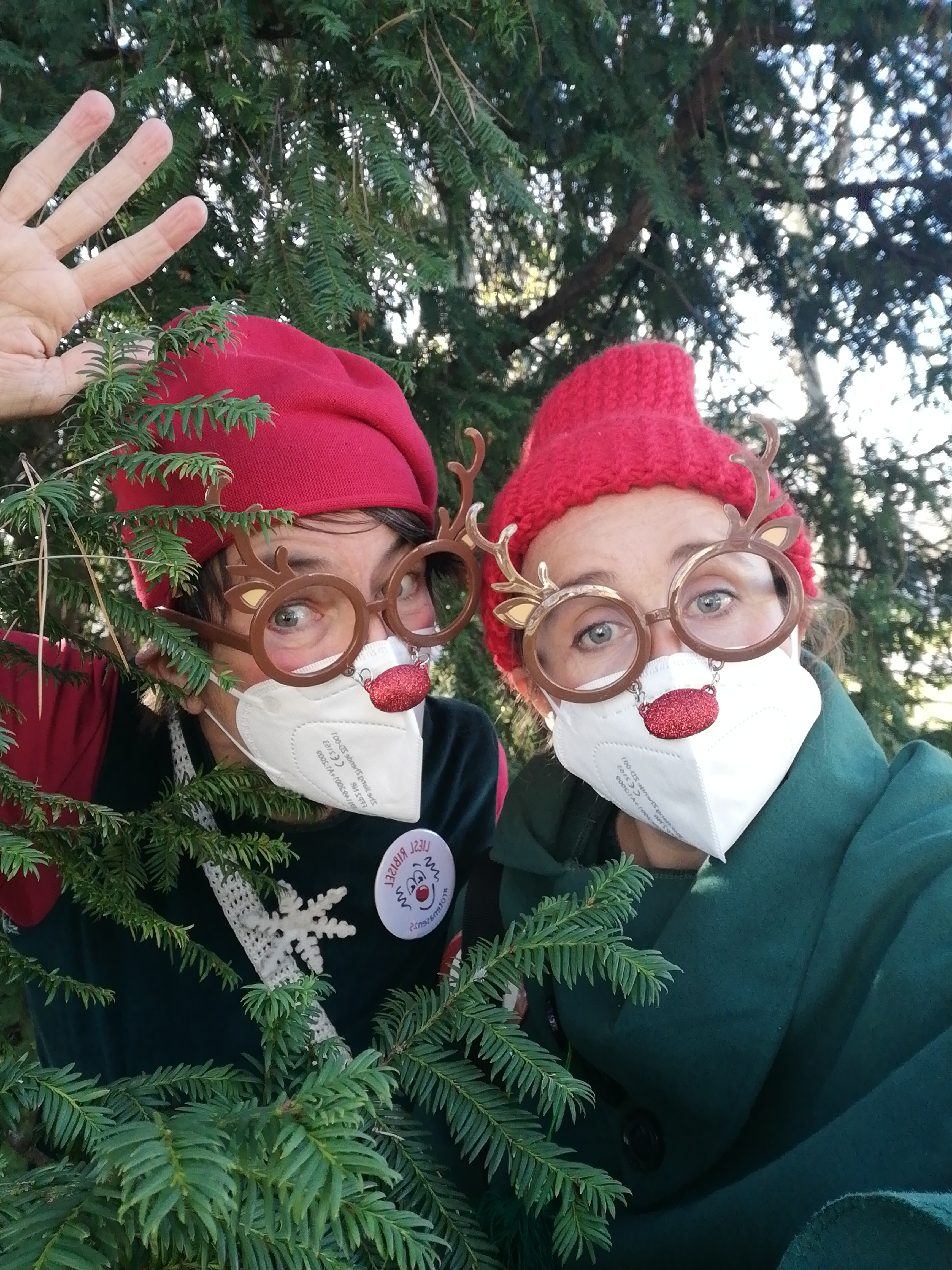 Zwei Clowninnen der ROTE NASEN mit Rentierbrille und roten Mützen stehe neben einem Tannenbaum.