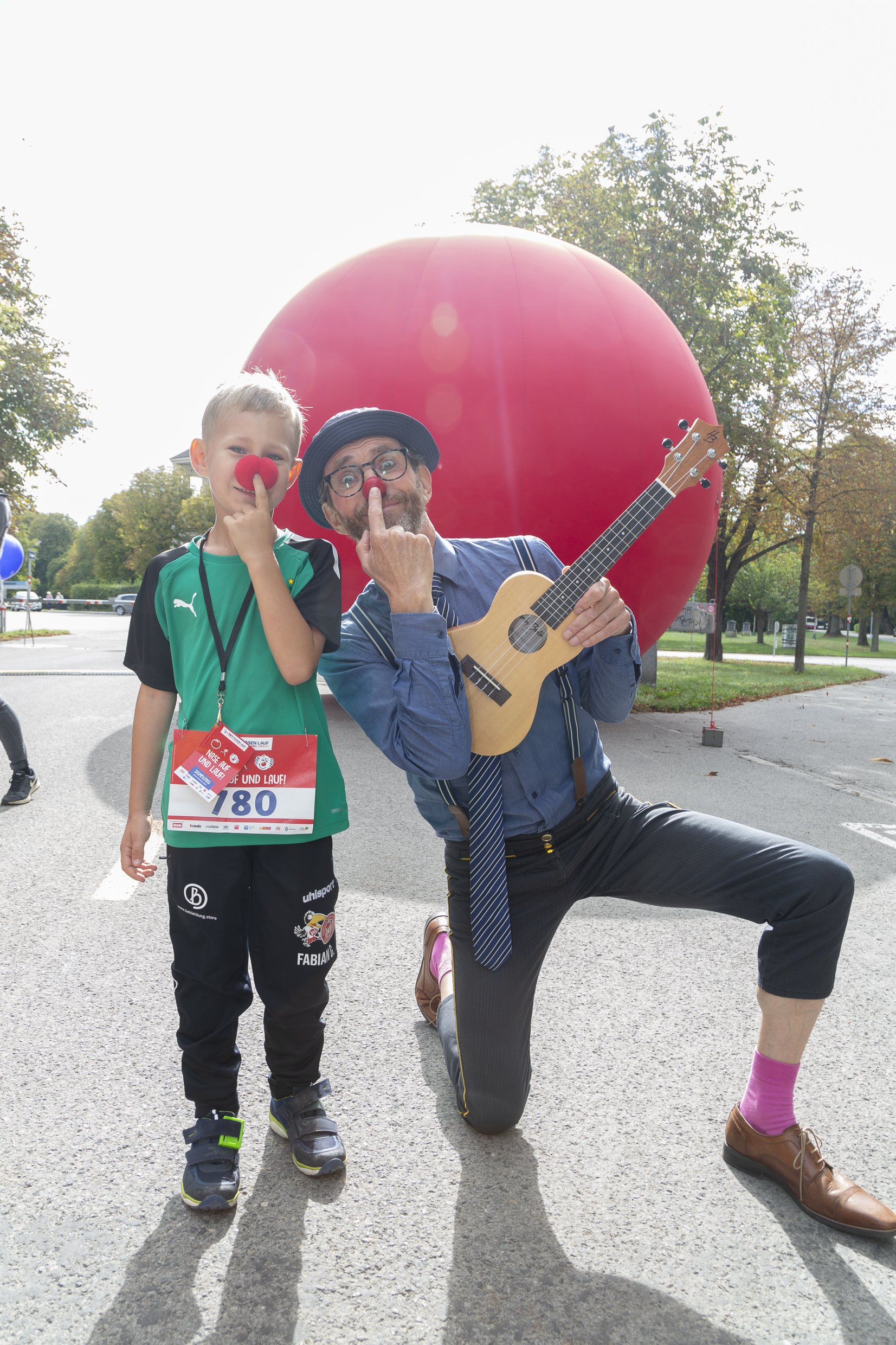 Ein ROTE NASEN-Clown mit Ukulele und ein kleiner Junge vor einer großen, roten Nase.