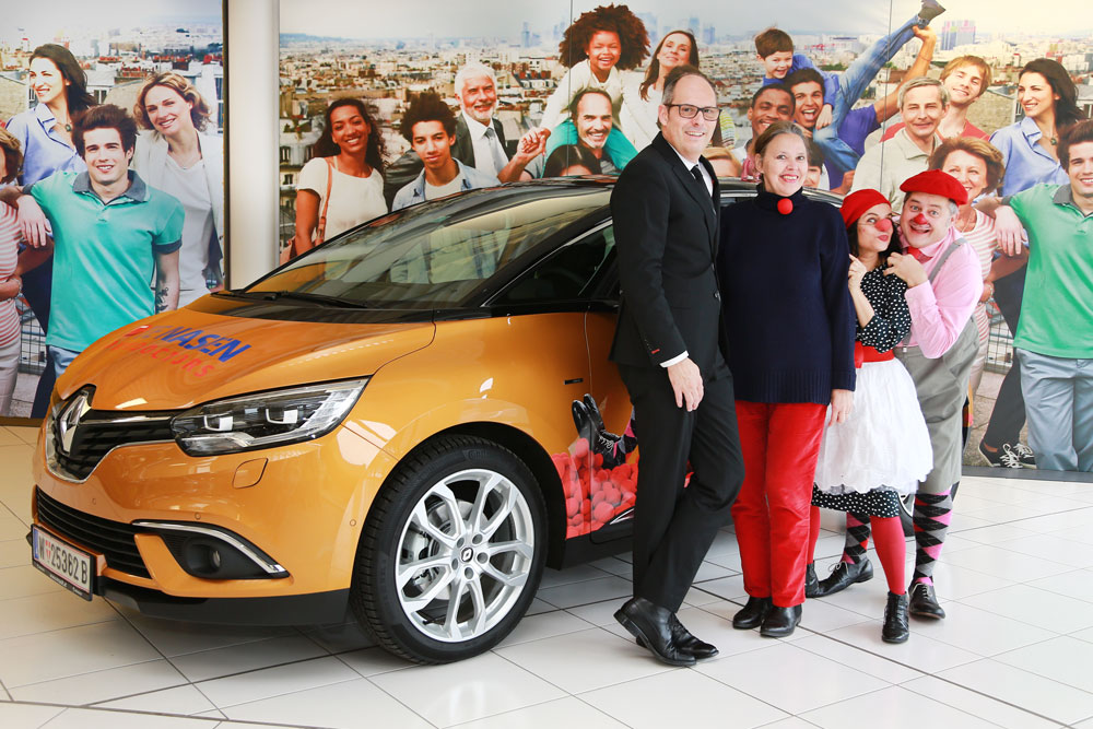 Ein Mann, eine Frau und zwei Clowns stehen vor einem orangene Auto