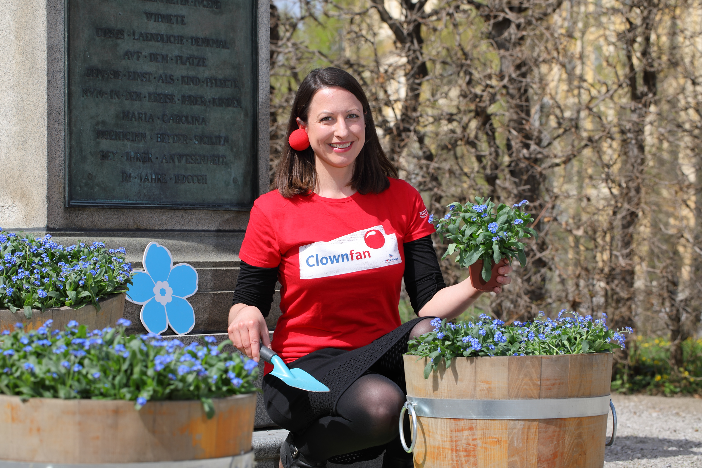 ROTE NASEN Mitarbeiterin posiert neben Blumentopf mit Vergissmeinnicht und pflanzt symbolisch Vergissmeinnicht