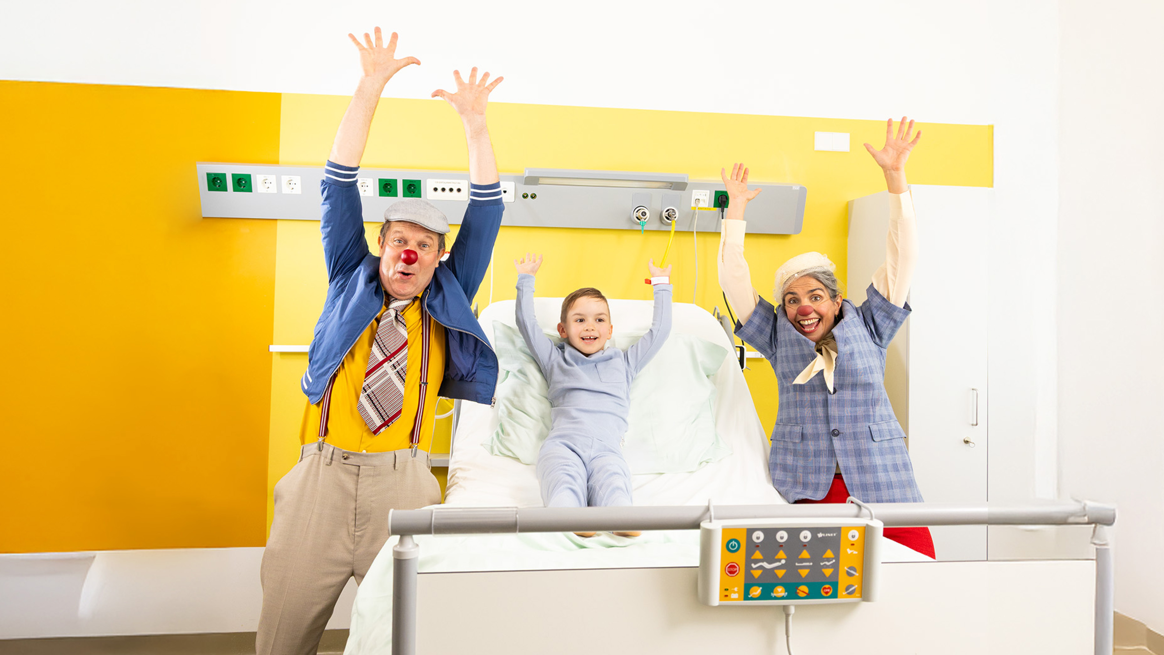 Zwei ROTE NASEN Clowns im Spital mit einem Kind und alle heben die Hände in die Luft.